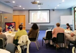 An education centre with PowerPoint presentation on screen. A number of people sit at round tables facing the screen with their backs to the camera.