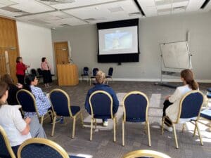 An education centre with presenter from a remote location on screen. There are chairs in the room with several healthcare professionals seated in them, their backs to the camera as they look at the screen.